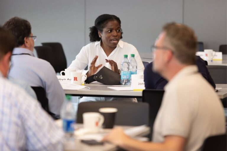 A woman in a white shirt engages in discussion with colleagues at a conference table during a meeting.