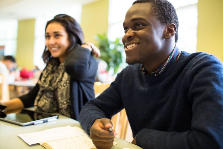 Two students sit together at a table, smiling and engaged, with notebooks and a tablet in front of them.
