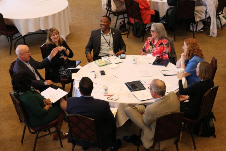 A group of professionals sits around a table in a conference setting, engaged in a lively discussion with papers, laptops, and coffee cups on the table.