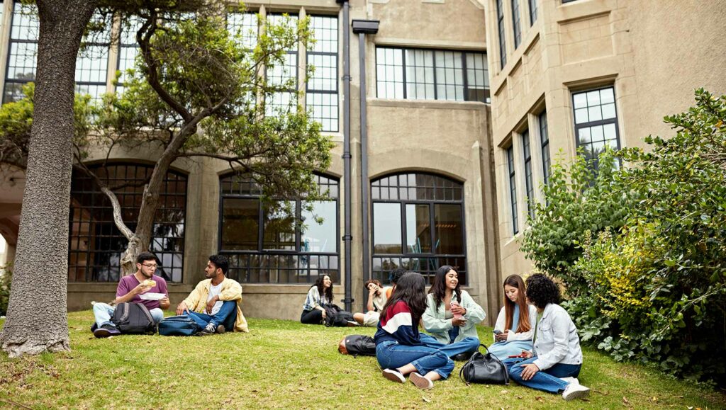 Students sit in small groups on a grassy lawn outside a large, historic building, talking and studying together.