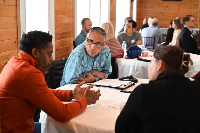 A group of people seated at tables, engaged in discussions with papers scattered around them in a well-lit room.